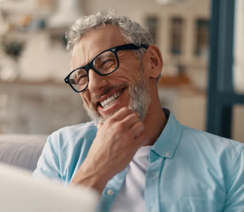 Smiling older man with gray hair and beard wearing black glasses and a light blue shirt, sitting indoors and looking at a screen.