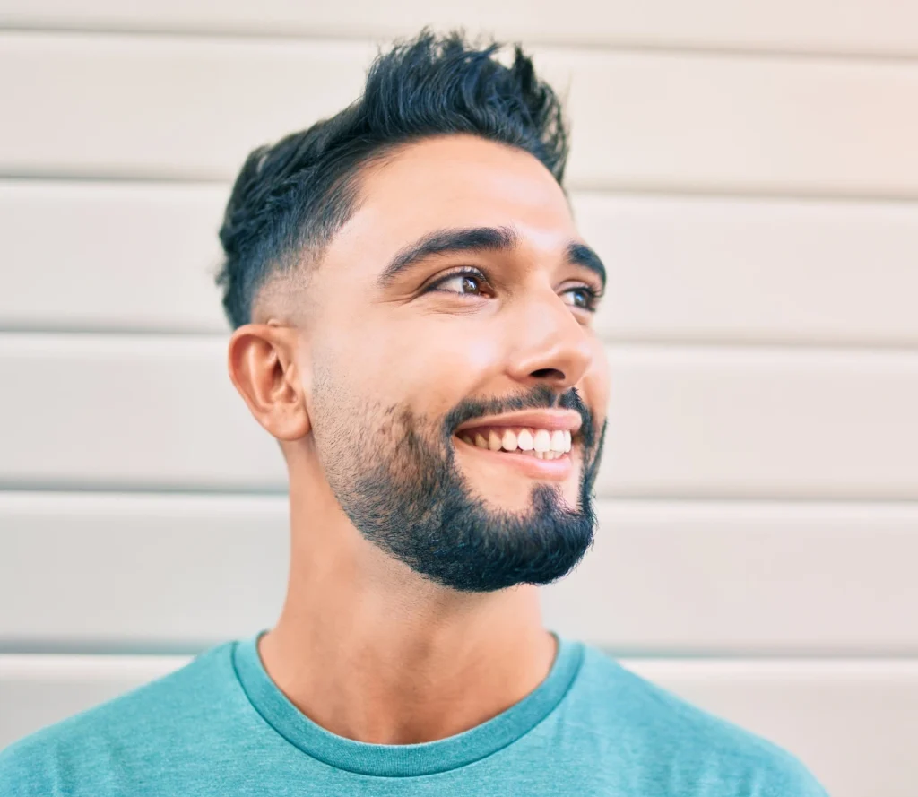 Smiling young man with a neatly groomed beard and short haircut looking to the side, wearing a teal shirt and standing in front of a light-colored wall with horizontal panels.