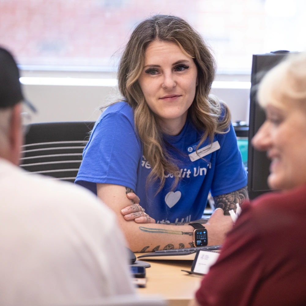 A credit union employee smiles while speaking with two customers at her desk.